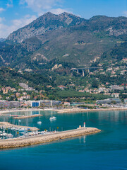 Fototapeta premium Aerial view of Menton, France shows the Old Port, marina, and turquoise bay with a stone breakwater and lighthouse, pastel town, viaduct, moored yachts, and midday light.