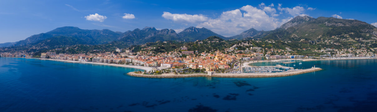 Aerial view of Menton on the French Riviera shows the Old Port, marina, promenade, and ochre bell towers by the Mediterranean and Alps, shot in clear daytime.