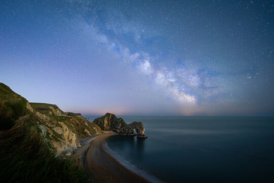 View of Durdle Door limestone arch under the Milky Way galaxy and starry night sky along the Jurassic Coast in West Lulworth, United Kingdom.