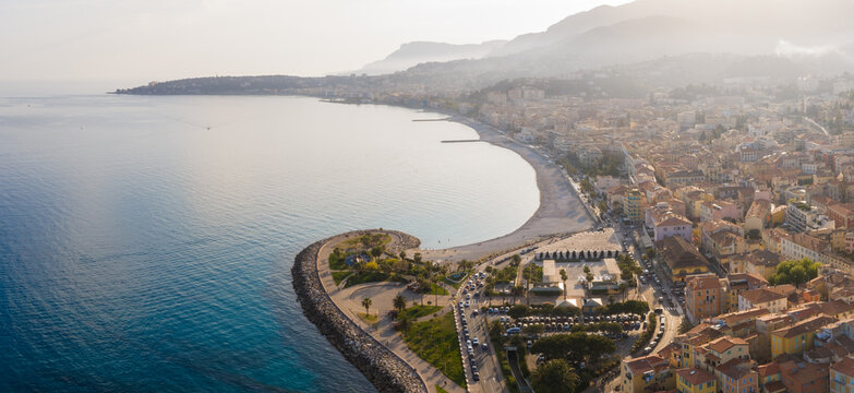 Aerial view of Menton, France shows Plage des Sablettes, stone jetty, pastel old town, promenade, gardens, docks, and hazy Alps Maritime foothills in warm late day light.