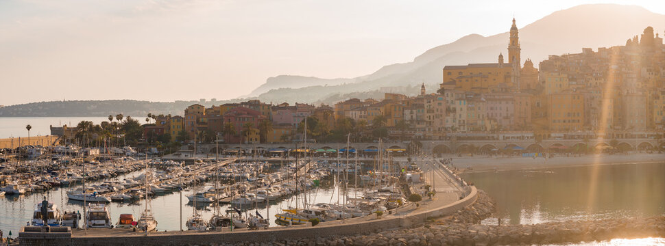 Aerial view of Menton, France shows the old town on the right, the marina of white hull boats along the breakwater, and the Basilica of Saint Michel by the layered hills.
