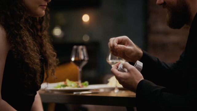 Side view cropped shot of bearded man in formalwear presenting diamond ring and proposing to happy surprised woman saying yes during romantic dinner in luxury restaurant