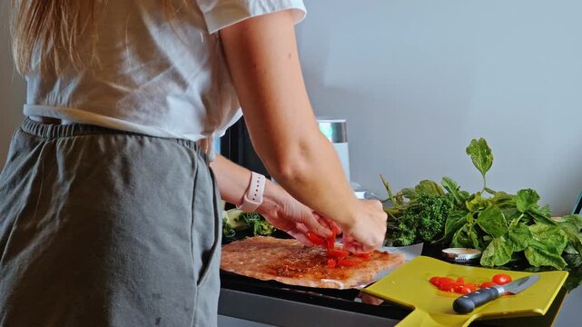 Woman placing sliced cherry tomatoes (Solanum lycopersicum) on raw ground chicken meat (Gallus gallus) in home kitchen, healthy meal preparation, culinary process.