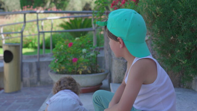 Toddler stumbling and falling while playing near older boy sitting on stone steps in garden with flowers and greenery in the background