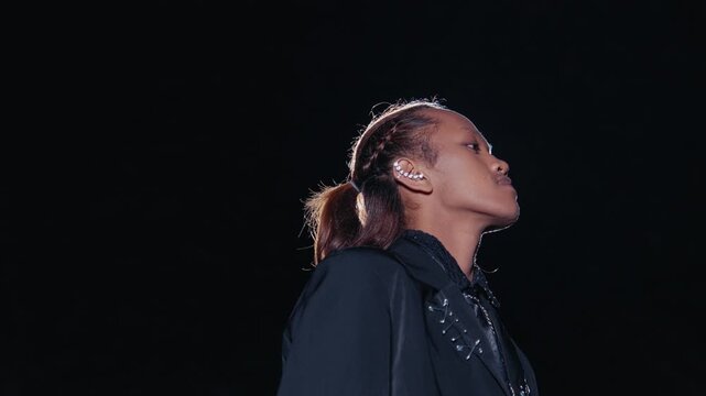An androgynous model with long hair in a ponytail and edgy fashion poses against a dark background, dramatically lit from behind by a bright spotlight. A cool and cinematic portrait.