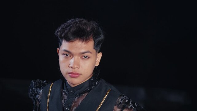 A portrait of a young, handsome man with a stylish haircut wearing an androgynous sheer blouse and vest. He poses with a confident and alluring gaze against a black background.