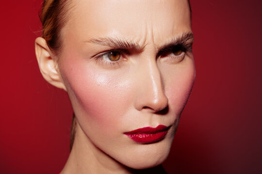 Close-up of a woman with red lipstick and flushed cheeks frowning against a red background. Concept of emotion, beauty, makeup, and facial expression in studio lighting.