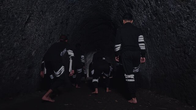 A group of men in matching outfits cautiously moves barefoot into a dark, ominous tunnel. Their low stances suggest a ritual, a performance, or a stealthy approach into the unknown.