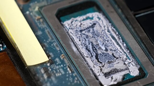 A macro view pulls back from a rectangular computer processor with old crusted thermal grease. The blue motherboard features memory chips and other electronic parts.