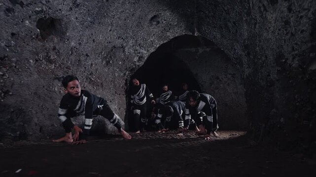 A group of male performers in matching outfits and glasses emerge from a dark cave, moving with intense, animal-like motions. A performance piece about struggle, emergence, or a primal tribe.