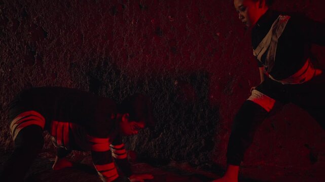A man and a woman in traditional costumes perform a tense, confrontational dance inside a dark cave. The moody red lighting enhances the feeling of a dramatic ritual, battle, or ancient story