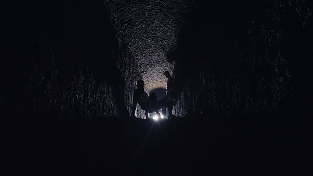 The silhouettes of several figures are backlit in a dark, narrow tunnel. One person lies on the ground while others stand over them, suggesting a mysterious sacrifice, rescue, or ancient ritual.