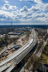 Obraz premium Aerial road and bridge works in Riga, Latvia, with machinery and crews. The Riga TV Tower rises on the horizon as clouds pass over leafless trees and low buildings.
