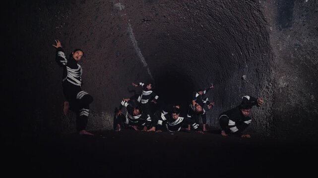 A theatrical performance unfolds as a group of dancers in skeleton-like outfits strike dramatic poses within a dark, cavernous space, led by a woman in a powerful stance.