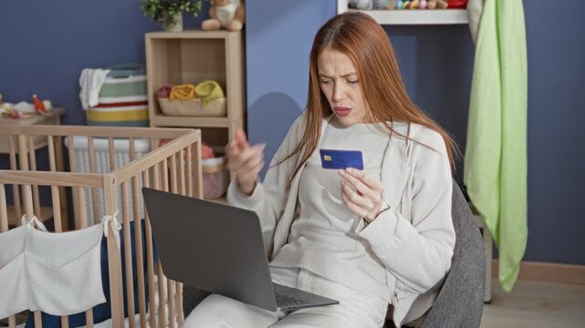Redhead woman holds a blue creditcard, points at a laptop and furrows her brow while seated beside a wooden crib in a nursery; financial stress.