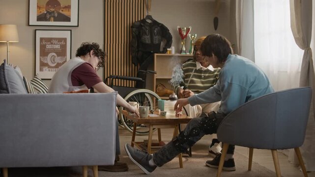 Three young adult men spending time together, playing poker and smoking hookah while having home party