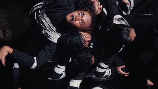 A high-angle, top-down view of a group of dancers with dramatic face paint, huddled together on the ground. Their slow, writhing movements create an abstract and intense image of unity or struggle.
