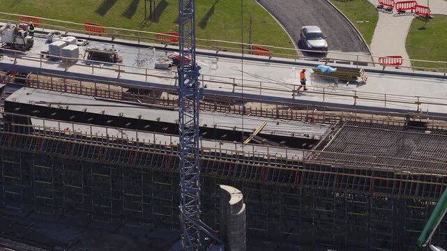 Aerial daytime view of Southern Bridge phase 4 in Riga, Latvia. Deep scaffold deck with rebar mats and formwork, tower crane mast, green pump arm, worker and barriers are present.