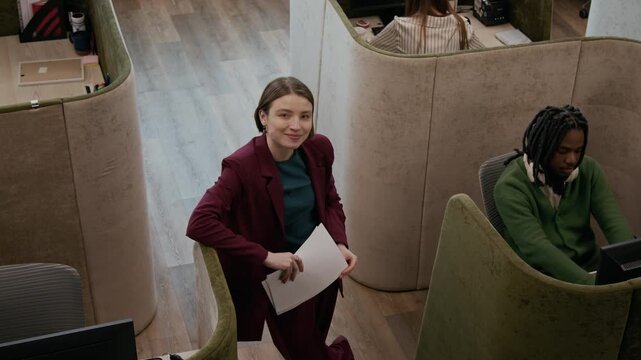 High angle portrait of young female manager holding documents and smiling confidently at camera while standing in modern open plan office with diverse employees working in private cubicles
