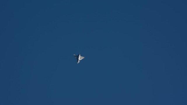 Military fighter jet in sharp banking roll at altitude against deep blue cloudless sky, delta wing silhouette visible from below, slow motion capture