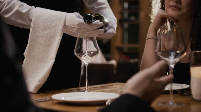 Cropped close up slowmo shot of hands of waiter wearing elegant white gloves pouring white wine into glass to unrecognizable couple enjoying romantic dinner in luxury restaurant