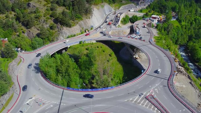 Drone footage captures the stunning aerial perspective of the bustling La Massana roundabout in Andorra, nestled within a picturesque mountain landscape. Cars navigate the multi-level intersection, sh