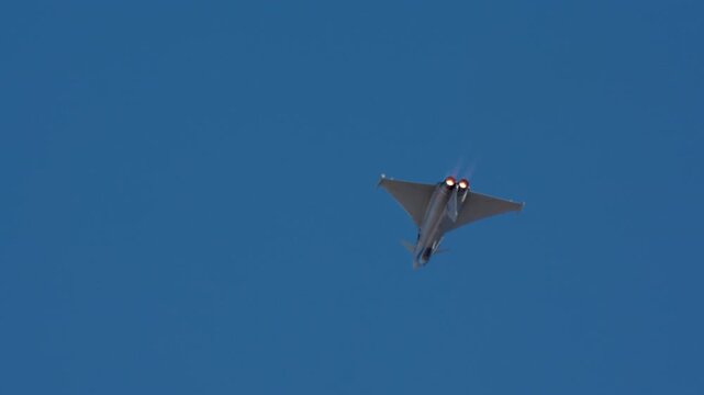 Military fighter jet seen from rear with two glowing afterburners firing against clear blue sky, slow motion capture
