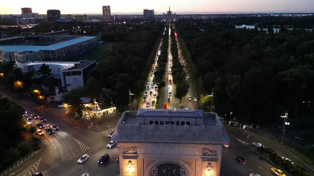 Nighttime aerial drone shot of the illuminated Arch of Triumph in Bucharest, Romania. View of the monument with city lights, traffic light trails, and evening urban atmosphere.