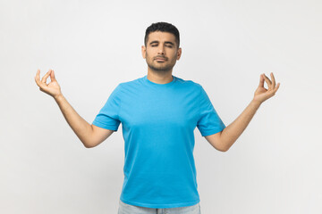 Fototapeta premium Portrait of relaxed young adult unshaven man wearing blue T- shirt standing practicing yoga, keeps eyes closed, trying to calm down. Indoor studio shot isolated on gray background.