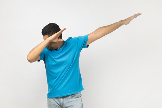 Portrait of positive excited unshaven man wearing blue T- shirt standing showing dab dance gesture, dancing, moving, internet trends. Indoor studio shot isolated on gray background.