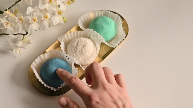 Hand gently pressing and touching colorful mochi ice cream desserts on a gold tray. Three Japanese rice cake sweets in blue, white coconut, and mint green colors displayed in paper cups with white orc