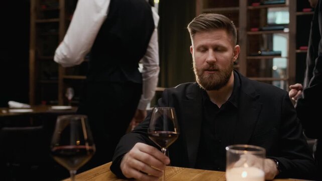 POV shot of young Caucasian man wearing suit talking while sitting at table in luxury restaurant holding glass of wine with couple of guests following waiter in blurred background