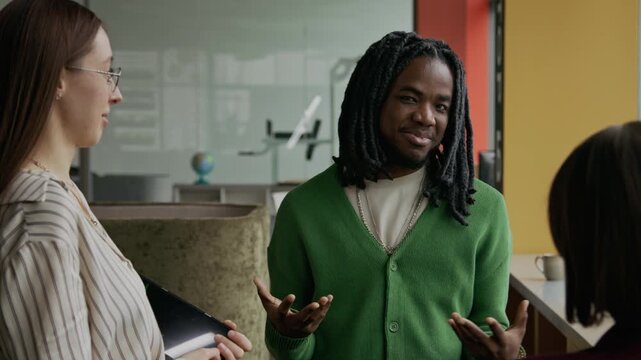 Young Black man with dreadlocks sharing his thoughts and explaining ideas about business project to two Caucasian female colleagues while working together in modern office