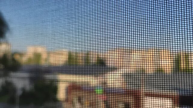 A close-up shot focuses on the mesh pattern of a mosquito screen on a window. In the background a blurry cityscape with apartment buildings is visible during a sunny day.