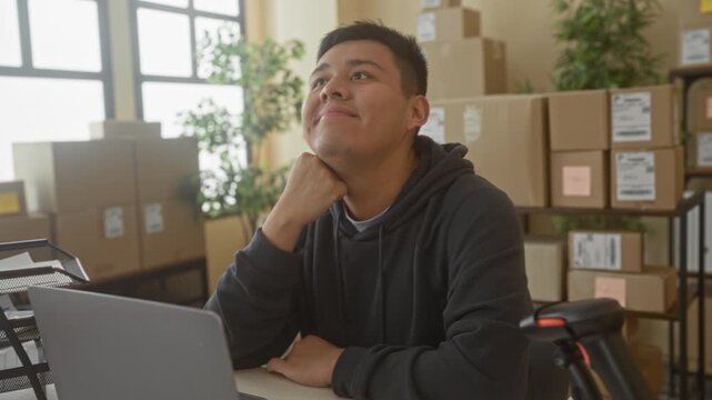 Man with laptop and parcels touching neck and resting chin on fist in building office; hopeful ambition.