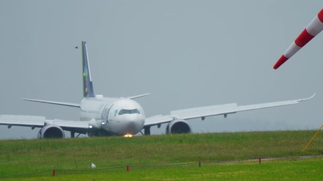 Wide-body commercial jet on airport taxiway facing camera, nose-on view, overcast sky, windsock visible, green grass in foreground, landing light on