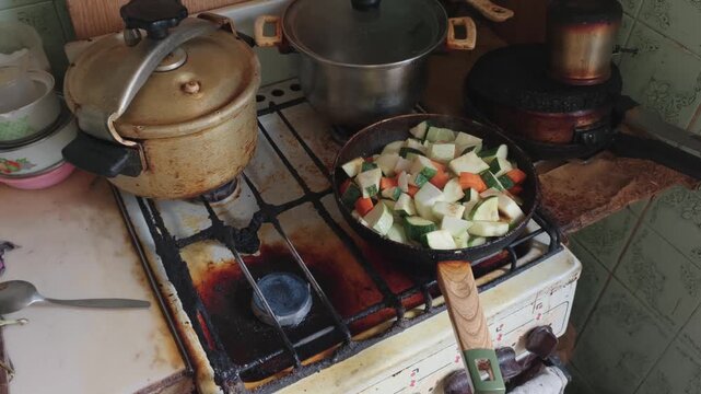 A frying pan with chopped zucchini and carrots sits on a very dirty gas stove. The messy greasy kitchen environment emphasizes concepts of neglect, poverty, and unsanitary living.