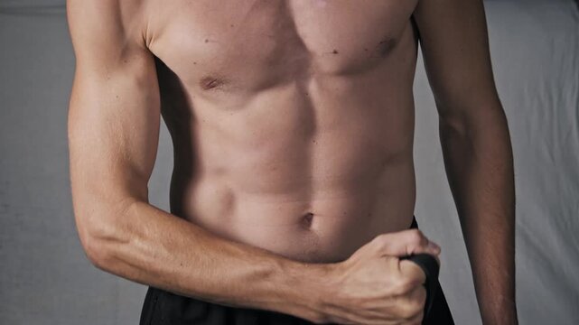 Close-up of a shirtless athletic man with a defined torso performing bicep curls. He uses a black resistance band to showcase muscle definition during strength training.