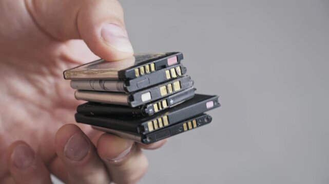 Side view of a hand holding a collection of used rechargeable lithium batteries. The pile of electronic waste symbolizes technology obsolescence and recycling needs.