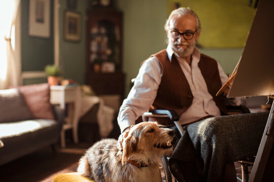 Senior man in wheelchair painting at home with dog