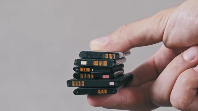 Side view of a hand holding a stack of discarded mobile phone batteries against a grey background. Focus on electronic waste disposal and environmental impact.