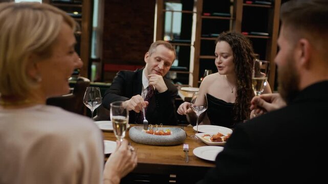 Over-the-shoulder handheld shot of adult Caucasian couples in elegant formalwear clinking glasses and enjoying appetizers at table in luxury restaurant with young sommelier pouring wine