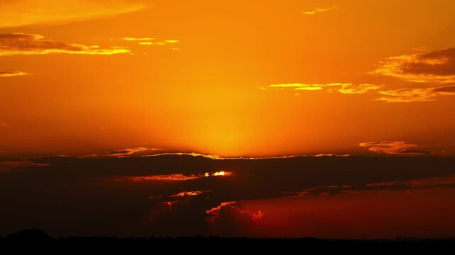 Timelapse of a vibrant orange sun as it disappears behind a thick layer of dark clouds. The scene casts a dramatic warm glow across the evening sky above a dark silhouetted horizon.