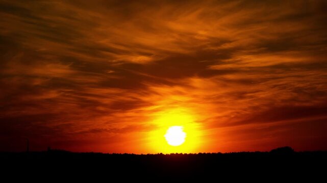 Time-lapse of a vibrant sun setting behind a silhouetted horizon with a fiery orange sky. Wispy clouds and a single bird fly past over a rural landscape in this scenic view.