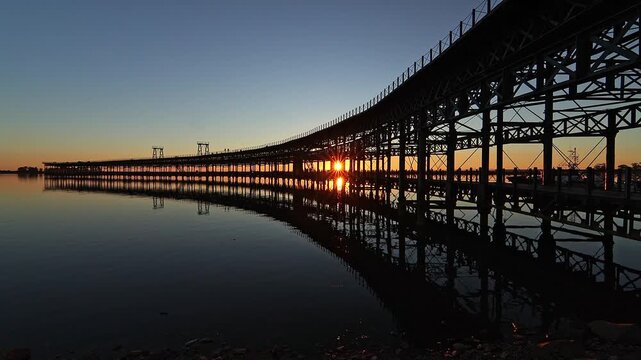 Wide shot of the historic iron pier in Huelva, featuring a vibrant sunstar through its structure and a perfect mirror reflection on the water. Great for heritage and travel documentaries.