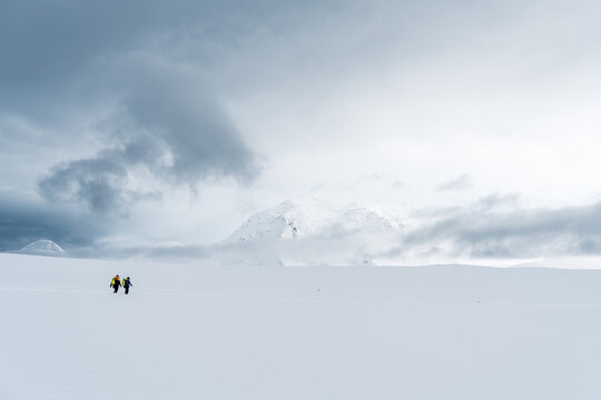 View of two explorers walking across a vast snowy plain towards a distant mountain peak under a dramatic cloudy sky in Antarctica.