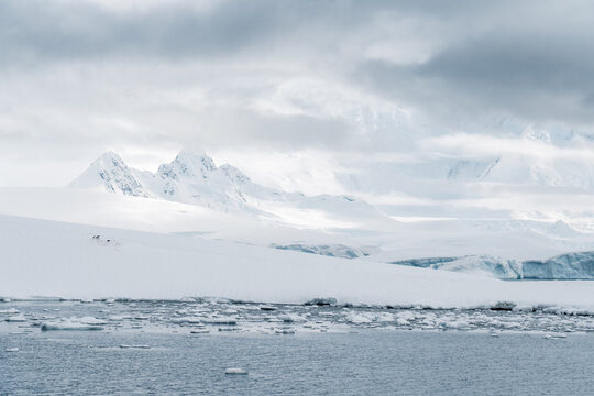 View of snow-covered mountain peaks and floating ice in the cold waters under a cloudy sky in Antarctica.