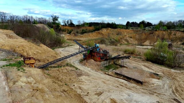 Aerial view of an industrial sand and gravel quarry, featuring heavy processing machinery with conveyor belts, sorted piles of extracted material, excavated landscape, and hilly terrain.