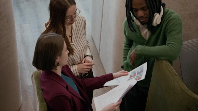 High angle shot of group of multiethnic gen Z coworkers standing in creative workspace with cubicles, reviewing business reports and discussing charts