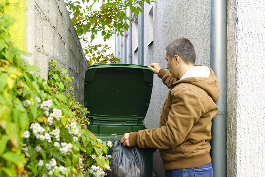Man lifting green wheelie bin lid to discard garbage bag in narrow passage. Portrays residential recycling behavior, waste interaction point, and interface between user and collection system.